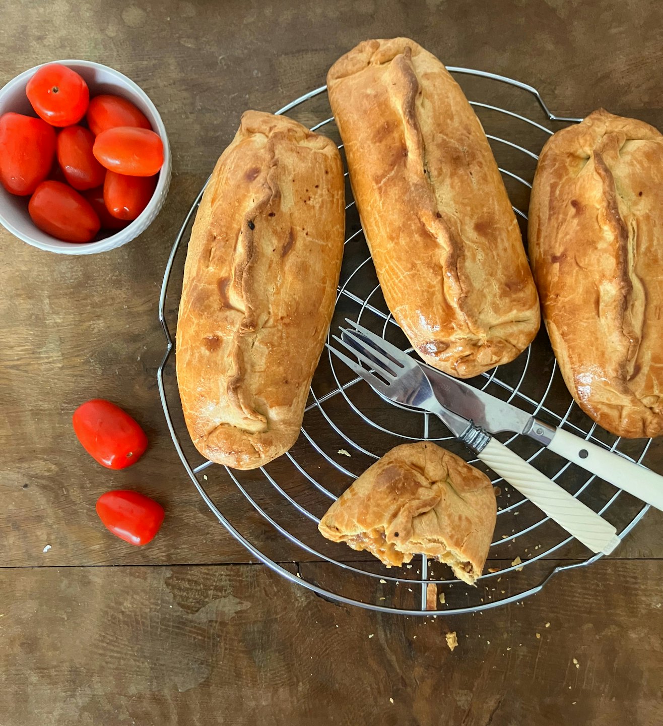Freshly baked pastries on a cooling rack with tomatoes and cutlery.
